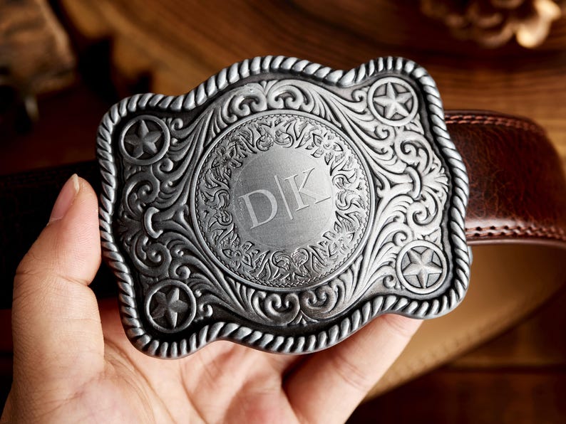 May include: Silver belt buckle with an ornate design featuring a circle with the initials "DK" in the center. The buckle has a rope-like border and three five-pointed stars.