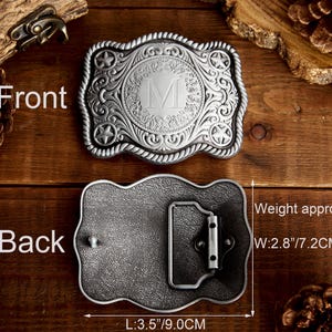 May include: Silver belt buckle with an ornate design featuring a rope border, a star on each side, and a circular center with a letter M. The back of the buckle is plain with a metal loop. The buckle measures 3.5 inches long and 2.8 inches wide and weighs 82 grams.