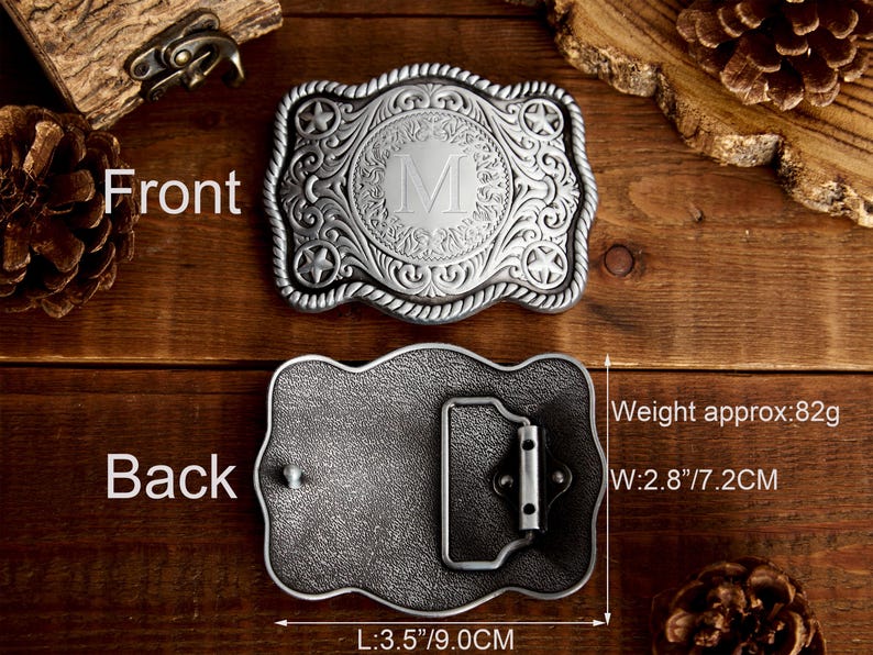 May include: Silver belt buckle with an ornate design featuring a rope border, a star on each side, and a circular center with a letter M. The back of the buckle is plain with a metal loop. The buckle measures 3.5 inches long and 2.8 inches wide and weighs 82 grams.