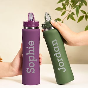 May include: Two personalized water bottles, one purple and one green, are held up. The purple bottle has the name "Sophie" in silver, and the green bottle has "Jordan". Both have a silver band and a flip-top lid.
