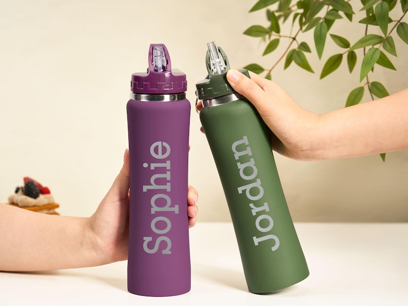 May include: Two personalized water bottles, one purple and one green, are held up. The purple bottle has the name "Sophie" in silver, and the green bottle has "Jordan". Both have a silver band and a flip-top lid.