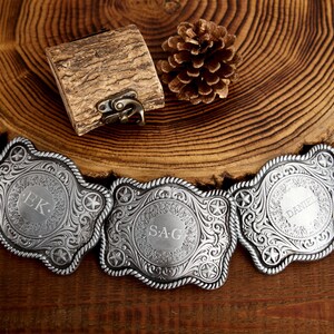 May include: Three silver belt buckles with ornate designs and engraved initials. The buckles are on a wooden surface.