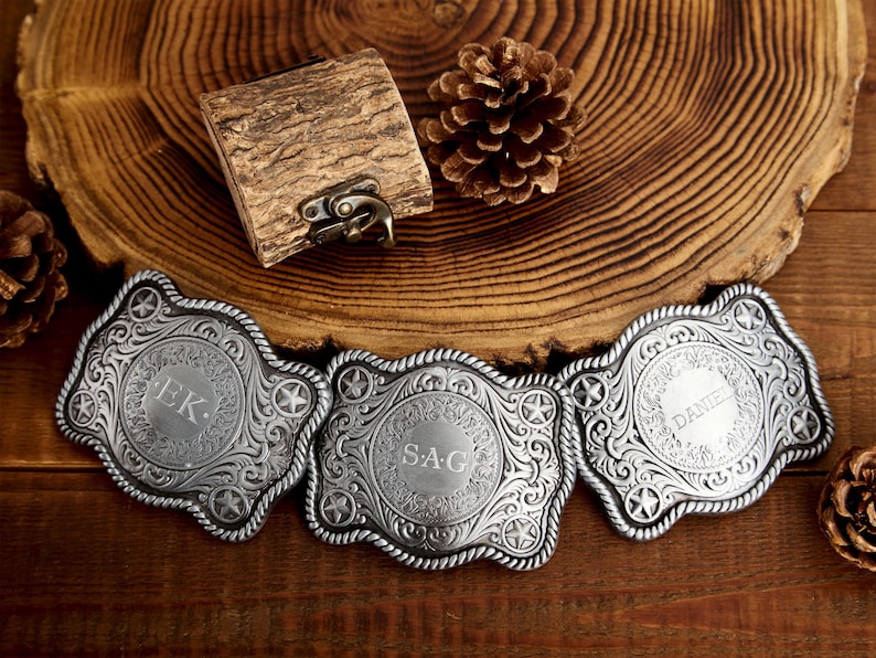 May include: Three silver belt buckles with ornate designs and engraved initials. The buckles are on a wooden surface.