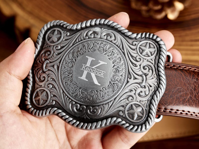 May include: Silver-toned belt buckle with an ornate design featuring a star and floral motifs. The buckle has a circular center with the initial "K" and the name "Kendrick" engraved.