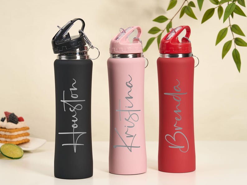 May include: Three personalized water bottles in black, pink, and red. Each bottle has a name in a cursive font: Houston, Kristina, and Brenda. The bottles have a flip-top lid and a silver ring for attaching to a bag.