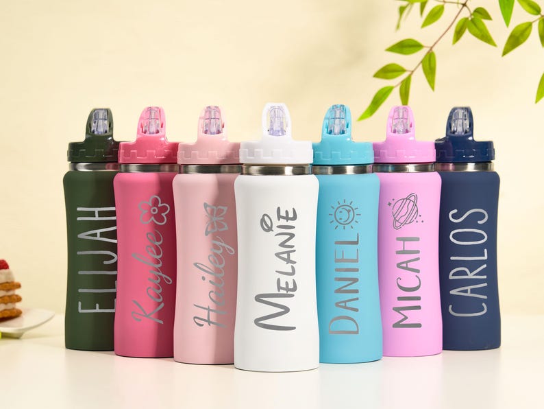 May include: A collection of personalized water bottles in various colors, including green, pink, light pink, white, blue, and navy. Each bottle features a name in a stylish font, with some also displaying decorative elements like a flower, butterfly, sun, and planet.