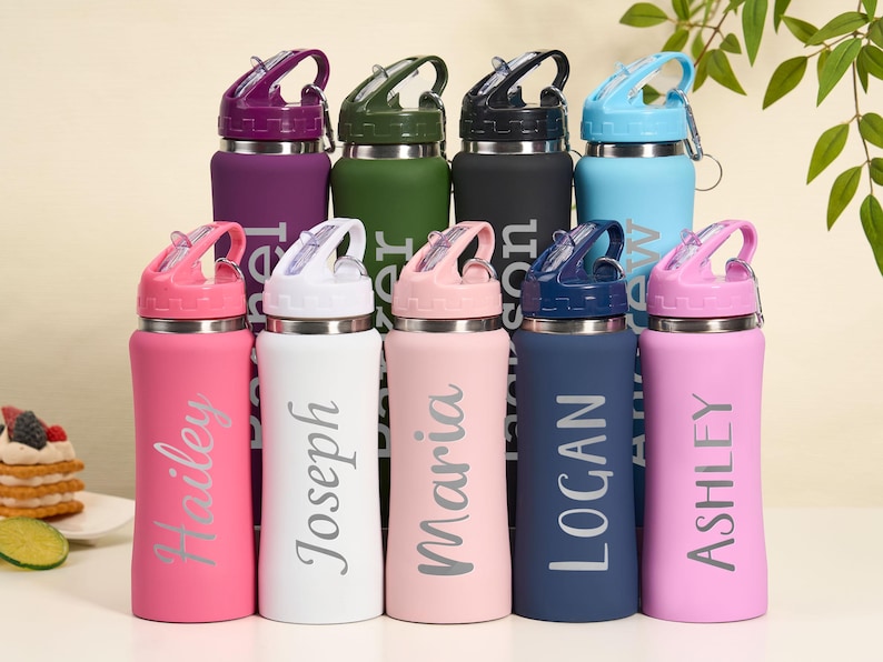May include: A collection of personalized water bottles in various colors, including pink, white, and blue. Each bottle has a flip-top lid and a name printed on the side. The bottles are designed for hydration and are suitable for everyday use.