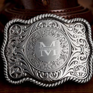 May include: Silver-toned metal belt buckle with an ornate, engraved design. The buckle features a central circular emblem with the letter "M" and the name "Maxwell". The edges are rope-like, and the corners have star-shaped accents. A Western-style accessory.