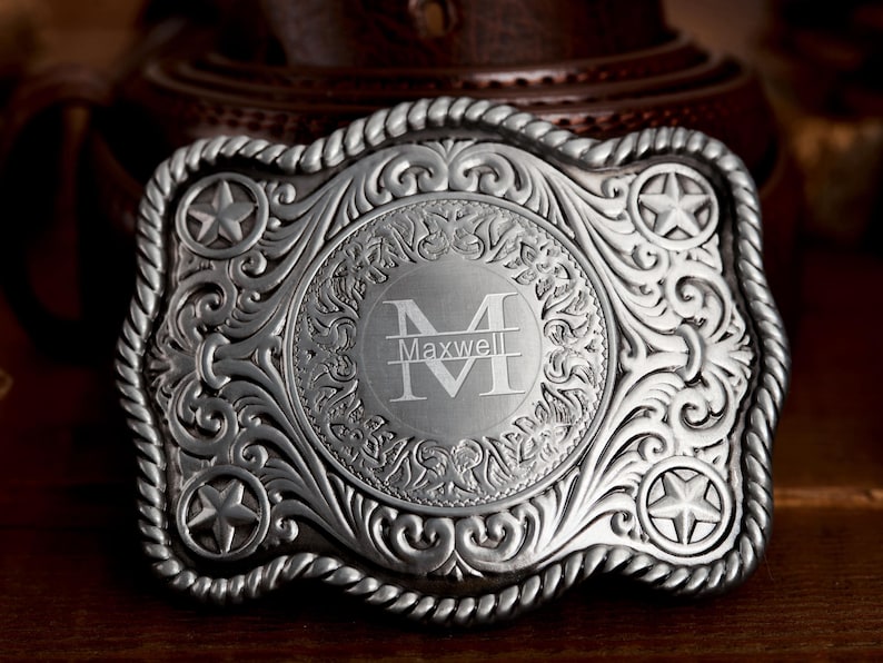 May include: Silver belt buckle with an ornate design featuring a star and a monogrammed initial "M" with the name "Maxwell" below.