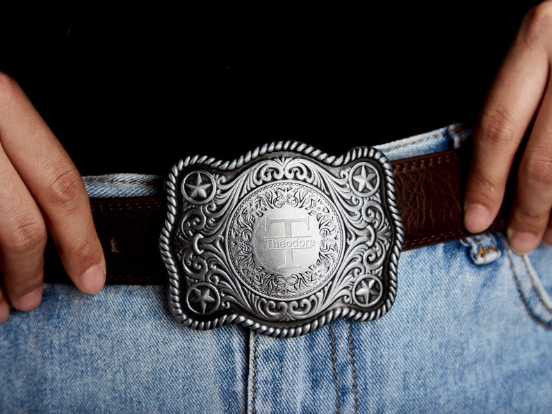 May include: A silver belt buckle with an ornate design and a central circle featuring the letter "T" and the name "Theodore".