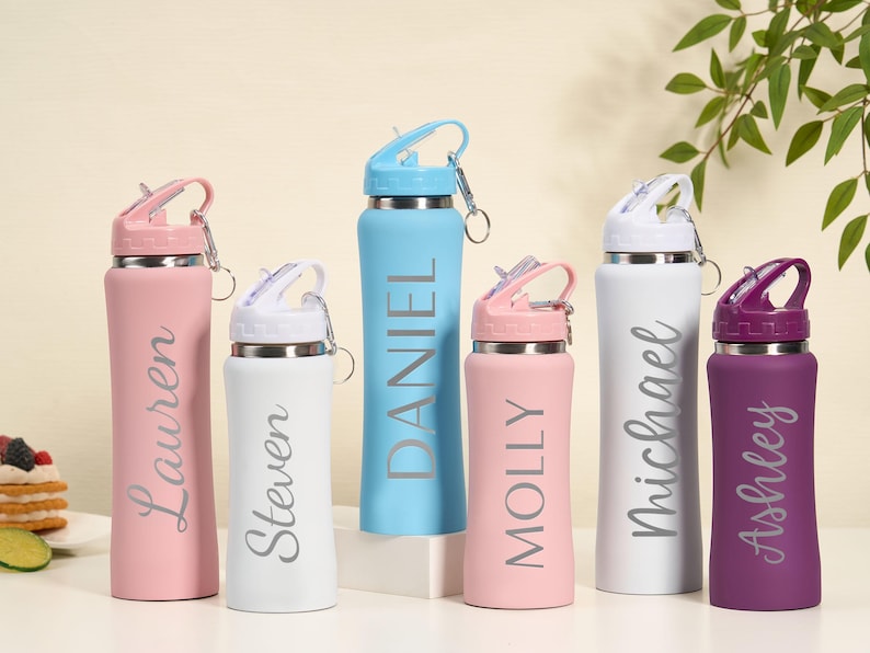 May include: A collection of personalized water bottles in various colors, including pink, white, blue, and purple. Each bottle features a name in a stylish font. The bottles have a flip-top lid and a silver carabiner clip.