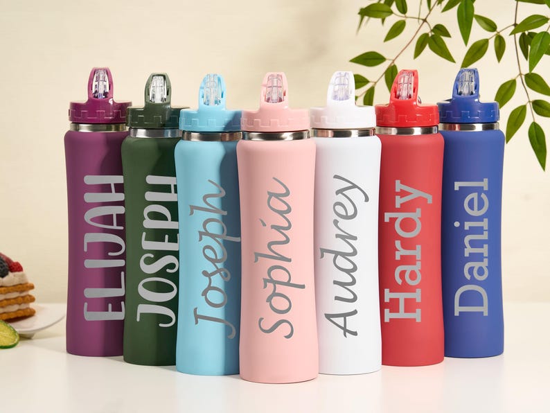 May include: A collection of personalized water bottles in various colors, including purple, green, blue, pink, white, red, and blue. Each bottle features a name in a stylish font. The bottles have a sleek design with a flip-top lid.
