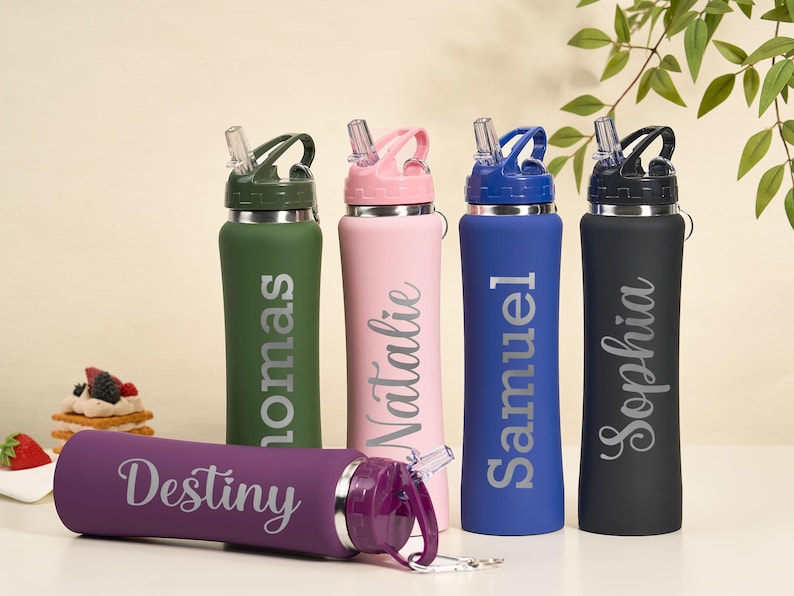 May include: A collection of personalized water bottles in various colors, including green, pink, blue, and black. Each bottle features a name in a stylish font. A purple bottle is lying down in front of the others. The bottles have a straw lid.