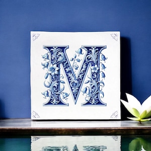 Op de afbeelding: Een witte keramische tegel met een blauw en wit bloemmotief met de letter "M".