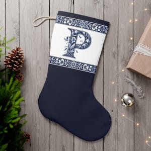 May include: A navy blue Christmas stocking with a white panel, floral design, and the letter "P" in blue. The stocking has a hanging loop. Festive decorations include a pine cone and bauble, creating a seasonal theme.