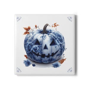 Op de afbeelding: Vierkante keramische tegel met een blauw-witte pompoen met een uitgesneden gezicht en bloemaccenten. De pompoen heeft een steel en is omgeven door bladeren en bloemen. De tegel heeft decoratieve hoekontwerpen.