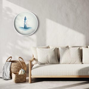 Op de afbeelding: Een witte wandklok met een blauw en wit aquarelontwerp van een vuurtoren op een golf. De klok hangt aan een witte muur boven een lichtbruine bank met witte kussens.