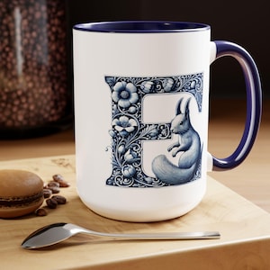May include: White ceramic mug with a blue rim and handle. The mug features a blue and white illustration of the letter "E" with a squirrel inside the letter. The illustration is surrounded by a floral design.