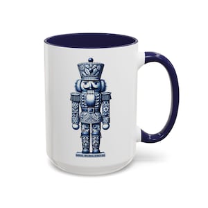 Op de afbeelding: Een witte keramische mok met een marineblauwe binnenkant en handvat. De mok heeft een gedetailleerde blauw-witte illustratie van een notenkrakersoldaat. Het ontwerp doet denken aan traditionele porseleinen patronen.