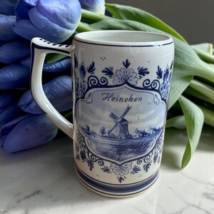 May include: A white ceramic mug with a blue and white windmill design and the word "Heineken". The mug has a curved handle and is decorated with floral patterns. Blue tulips are in the background.