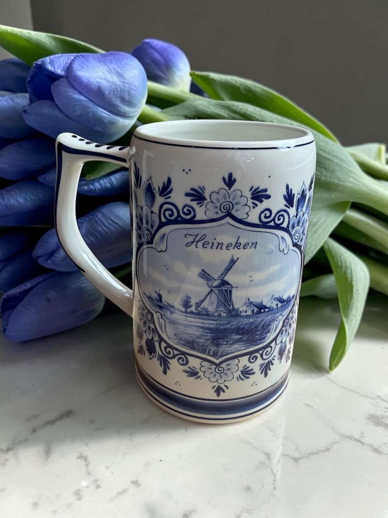 May include: A white ceramic mug with a blue and white windmill design and the word "Heineken". The mug has a curved handle and is decorated with floral patterns. Blue tulips are in the background.
