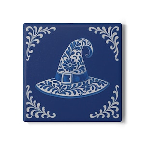 Op de afbeelding: Vierkante, diepblauwe decoratieve tegel met een wit en blauw heksenhoedontwerp. De hoed is versierd met bloempatronen en een gesp. Witte bloemenhoekaccenten omlijsten de hoed en creëren een grillige, Halloween-thema-esthetiek.