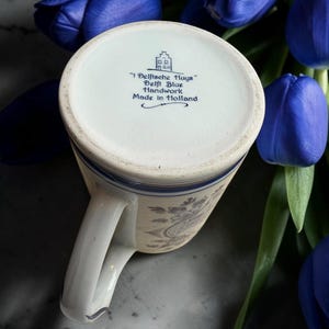May include: A white and blue Delft Blue handwork mug with a handle, featuring floral patterns. The bottom of the mug has text that reads "Delftsche Huys" and "Made in Holland". The mug is surrounded by blue tulips.