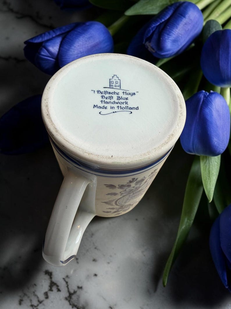 May include: A white and blue Delft Blue handwork mug with a handle, featuring floral patterns. The bottom of the mug has text that reads "Delftsche Huys" and "Made in Holland". The mug is surrounded by blue tulips.