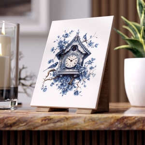 May include: A white ceramic tile with a blue and white illustration of a cuckoo clock with birds perched on branches. The clock face has Roman numerals and the clock is surrounded by blue flowers.