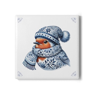 Op de afbeelding: Vierkante keramische tegel met een gedetailleerde illustratie van een roodborstje. De vogel draagt een blauw-wit patroonmuts en sjaal. De tegel heeft een witte achtergrond met blauwe decoratieve hoekaccenten.