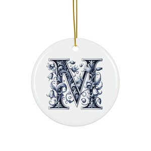 Op de afbeelding: Een ronde, witte keramische ornament met een gouden ophangtouwtje. Het ornament toont een gedetailleerd, blauw bloemenontwerp in de vorm van de letter "M". Het ontwerp is geplaatst tegen een witte achtergrond. Een decoratief item.