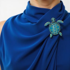 Könnte beinhalten: Eine blau-goldene Schildkrötenbrosche mit türkisfarbenen Akzenten. Die Brosche ist an einem drapierten blauen Stoff befestigt. Die Schildkröte hat eine goldene Kontur, einen türkisfarbenen Körper und türkisfarbene Akzente an Kopf und Flossen.