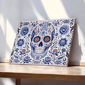 Może przedstawiać: Biały wydruk na płótnie z niebieskim i pomarańczowym wzorem czaszki cukrowej. Czaszka jest otoczona niebieskimi i białymi wzorami kwiatowymi. Tekst "Dia de los Muertos" znajduje się na górze wydruku.