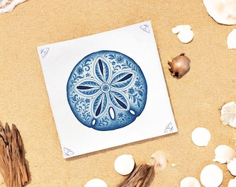Puede incluir: Una baldosa cuadrada y blanca con un diseño detallado de dólar de arena azul. El dólar de arena está rodeado de patrones florales, sobre un fondo de playa arenosa con conchas marinas y madera flotante.
