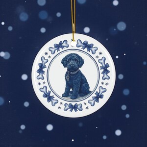 Op de afbeelding: Een ronde, witte keramische ornament met een illustratie van een zwarte hond. De hond draagt een halsband en is omgeven door een blauw-witte rand van strikken, botten en sneeuwvlokken. Een gouden touwtje is bevestigd om op te hangen.