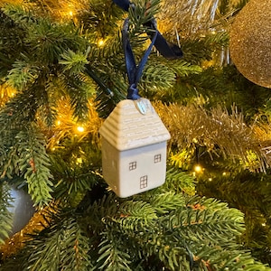 Op de afbeelding: Een keramische huisornament voor de kerstboom met een beige dak en witte muren, met drie kleine ramen. Het hangt aan een donkerblauw lint in een kerstboom met gouden slingers en lichtjes. Een gouden bal is ook zichtbaar.