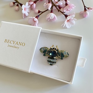 Peut inclure: Broche abeille dorée ornée de cristaux verts, bleus et noirs, présentée dans un écrin blanc. La broche comporte un grand cristal bleu foncé au centre. La boîte porte l'inscription "BECYANO Jewellery". Des fleurs de cerisier roses en arrière-plan.