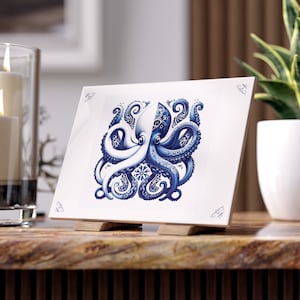 Op de afbeelding: Vierkante witte keramische tegel met een blauw-wit octopusontwerp. De octopus heeft ingewikkelde patronen en wordt op een houten standaard gepresenteerd. Decoratieve accenten bevinden zich in elke hoek.