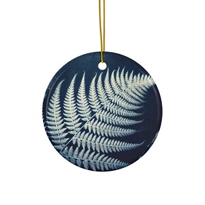 Op de afbeelding: Een ronde kerstbal van keramiek met een witte varenblad op een donkerblauwe achtergrond. De kerstbal heeft een gouden koord om op te hangen.