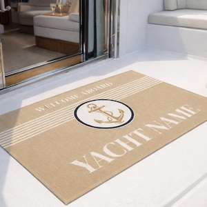 Op de afbeelding: Beige rechthoekige welkomstmat met de woorden "WELCOME ABOARD" en "YACHT NAME" in het wit. Een marineblauwe en witte cirkel met een gouden anker staat in het midden. De mat heeft witte strepen.