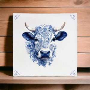 Könnte beinhalten: Quadratische Leinwandkunst mit einer detaillierten Illustration eines Kuhkopfes. Die Kuh ist mit blauen floralen Mustern und Akzenten verziert, vor einem weißen Hintergrund. Das Kunstwerk hat dekorative blaue Eckdesigns und wird auf einer Holzoberfläche präsentiert.