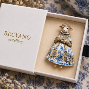 Peut inclure: Une broche dorée en forme de mouton portant une robe à motif floral bleu et blanc, ornée de petits cristaux bleus et transparents. La broche est présentée dans une boîte blanche avec le texte BECYANO Jewellery imprimé en lettres dorées.
