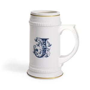 Op de afbeelding: Witte keramische bierpul met een gouden rand en handvat. De pul heeft een blauw bloemmotief met de letter "J". Het ontwerp is gedetailleerd en sierlijk, met een klassieke, vintage esthetiek.