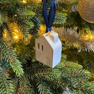 Op de afbeelding: Een wit keramisch huisornament met een donkerblauw lint dat aan een kerstboom hangt. Het huis heeft een schoorsteen en twee ramen. Een goudkleurig label is aan het lint bevestigd. De achtergrond bevat andere ornamenten en dennentakken.