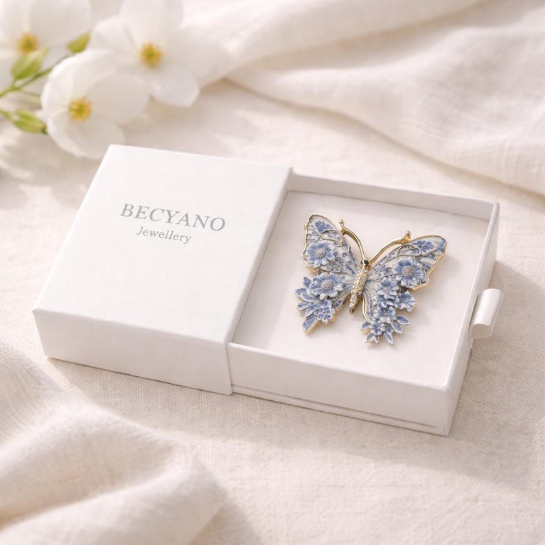 May include: A butterfly brooch with a gold-tone frame and detailed blue floral design, displayed in an open white box. The box features the brand name "BECYANO Jewellery". The brooch is a delicate piece of jewellery.
