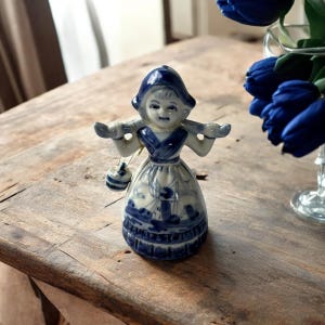 Op de afbeelding: Een blauw en wit porseleinen beeldje van een Nederlands meisje met een mandje. Het beeldje draagt een traditionele Nederlandse muts en jurk. De jurk heeft een gedetailleerde blauw-witte nautische scène. Het beeldje is ongeveer 10 cm hoog.