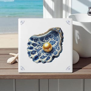 May include: Square ceramic tile featuring a detailed illustration of an oyster shell with a golden pearl. The shell is decorated with blue floral patterns and a gold trim. The tile has small blue corner accents.