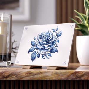 Op de afbeelding: Een witte keramische tegel met een blauw en wit bloemenmotief. De tegel heeft een enkele roos met ingewikkelde details en bladeren. De tegel staat op een houten standaard.