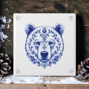 May include: A square white ceramic tile featuring a detailed blue ink illustration of a bear head. The bear is decorated with intricate floral and foliate scrollwork patterns. Small blue decorative corner accents are present on the tile.