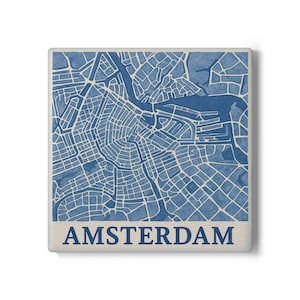 Op de afbeelding: Vierkante tegel met een blauw-witte kaart van Amsterdam. De kaart toont de straten en waterwegen van de stad. Het woord "AMSTERDAM" staat in blauw onderaan. De tegel heeft een neutrale rand.